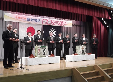 新春のつどいと書かれた横断幕があるステージに、2個の酒樽があり、その後ろに市長の他、関係者9人が枡を持って並び、その中の1人があいさつをしている写真