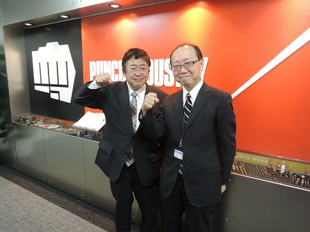 山本市長と武田雅亮社長がグーのイラストがある会社の看板前でガッツポーズをとっている写真