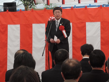 紅白の幕を背景に赤の胸章をつけ、あいさつを述べている山本市長と それを座って聞いている多くの参加者の方々の写真