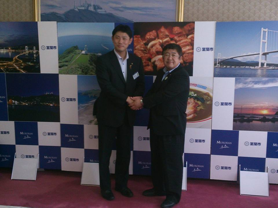 室蘭市のバックパネルの前で両手で握手をする青山室蘭市長と山本市長の記念写真