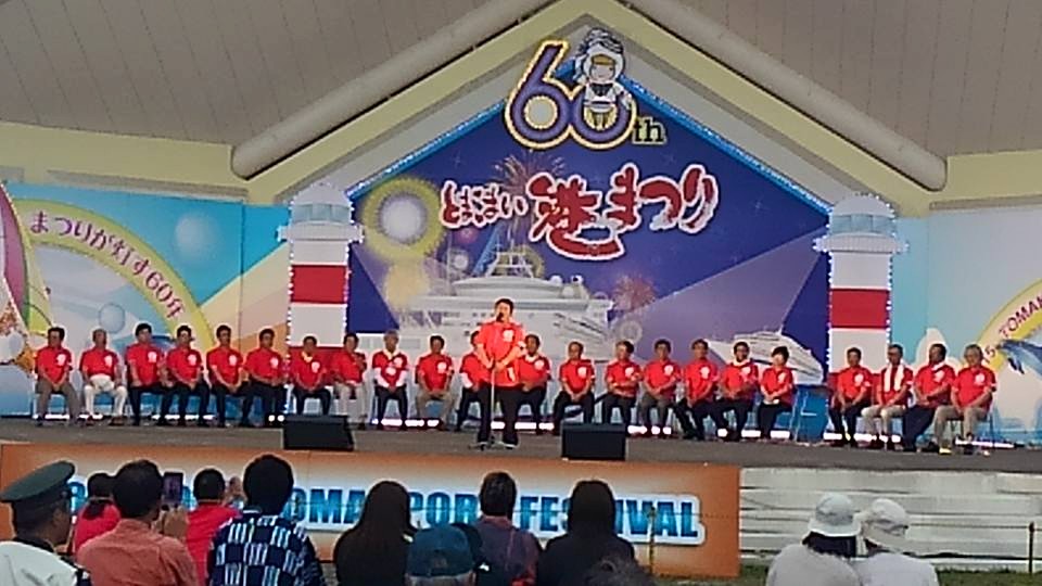 「とまこまい港まつり」と書かれた看板などで装飾された舞台上の椅子に座り横一列に並ぶ赤いTシャツを着た関係者の方々の前で、スタンドマイクに向かい話をする市長の写真