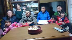 自宅のコタツのある部屋の中央に座る藤田スエノさんと市長が横に並び、4名のひ孫やご家族に囲まれた記念写真