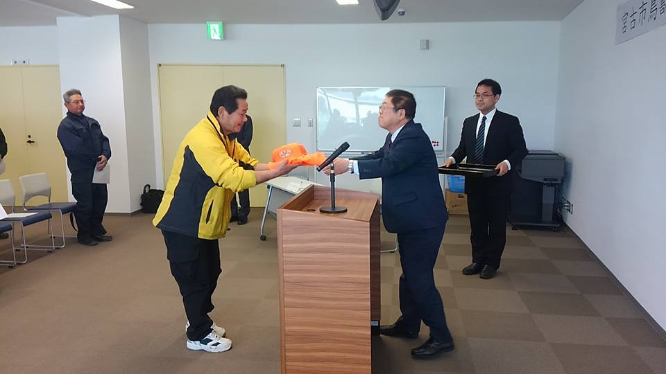檀上を挟んで黄色のジャンパーを着た隊員と市長が向かい合って立ち、市長からオレンジ色のキャップとジャンパーを受け取っている様子の写真