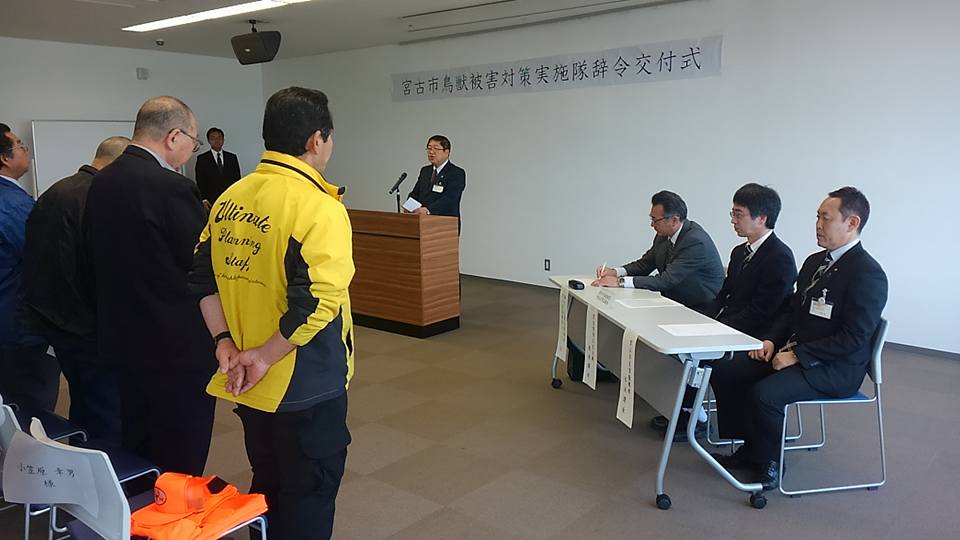「鳥獣被害対策実施隊辞令交付式」と書かれた横幕が壁の上に貼られた室内で、自席から立ち上がった隊員が、檀上に立つ市長の話を聞いている交付式の様子の写真