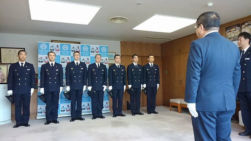 バックパネルが設置された室内で、紺色の制服に白の手袋をはめ横一列に並んだ7名の消防職員と市長が向かい合って立っている写真