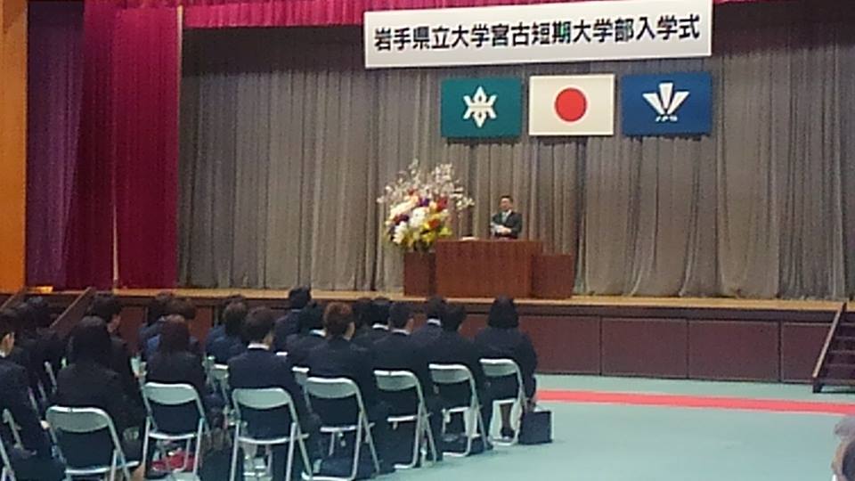 「岩手県立大学宮古短期大学部入学式」と書かれた横断幕、国旗と県旗に校旗の3つの旗が飾られたステージの檀上に立つ市長の話を、体育館に設置された椅子に座っている新入生が聞いている岩手県立大学宮古短期大学部の入学式の様子の写真
