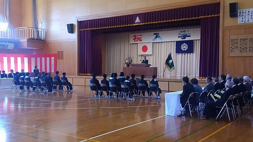 ステージ上に「祝 入学」と書かれた横断幕、国旗と市旗の2つの旗が飾られたステージの檀上に立つ市長の話を、体育館の左側と右側に座った関係者の方々、中央に並んで座った新入生が聞いている、新里中学校の入学式の様子の写真