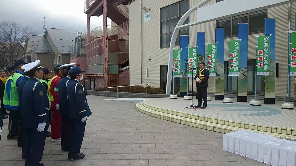 「交通安全運動実施中」と書かれたのぼり旗が並んで設置された屋外広場で、整列した警察官や関係者と向かい合って立つ市長がマイクスタンドの前で話をしている写真
