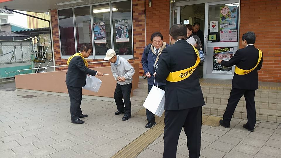 スーツの上に黄色のタスキを掛けた市長と2名の関係者が、レンガ調の外観の建物から出てきた方達に啓発グッズを手渡している交通安全運動の様子の写真