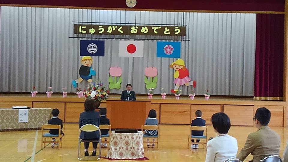 ステージ上に「にゅうがく おめでとう」と書かれた横断幕、国旗と市旗に校旗の3つの旗、チューリップ花や子どもが走っているイラストが飾られ、体育館の中央に4名の新入生と先生、右後方に保護者が座り、檀上で挨拶を行っている市長の話を参加者が聞いている川井小学校の入学式の様子の写真