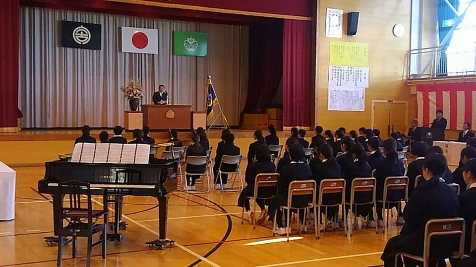 紅白幕が貼られた体育館にピアノが設置され、国旗と市旗、校旗が飾られたステージの檀上に立つ市長の話を並んで座った新入生が聞いている、崎山中学校の入学式の様子の写真