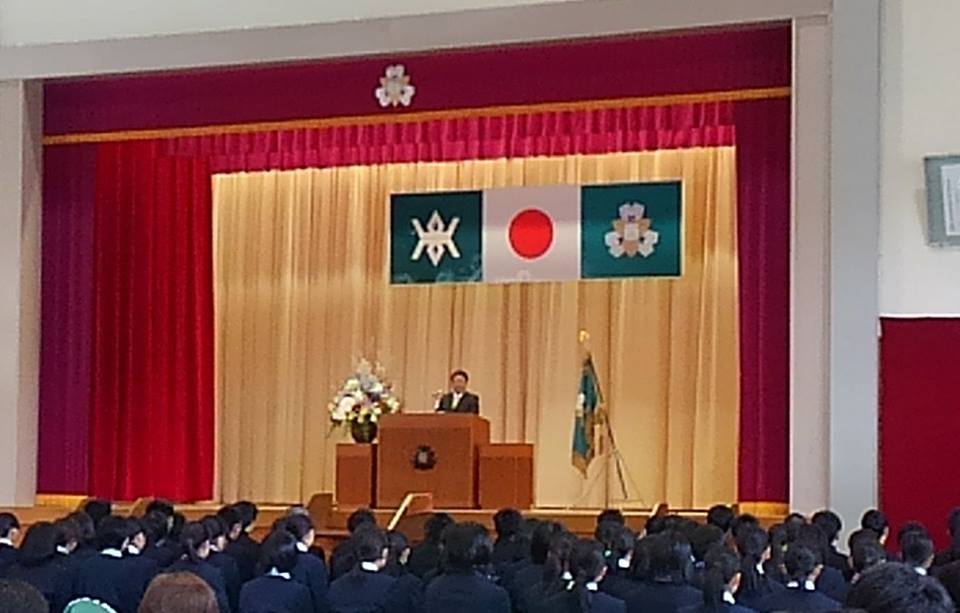 体育館に新入生が並んで座り、国旗と県旗に校旗の3つの旗が飾られたステージの檀上に立つ市長の話を聞いている宮古商業高等学校入学式の様子の写真