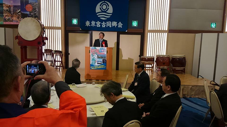 小さなステージ上に「東京宮古同郷会」と書かれた旗が飾られ、円卓の席に座っている参加者が檀上に立つ市長の話を聞いている写真