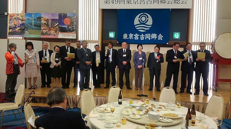 「第49回東京宮古同郷会総会」と書かれた横断幕と旗が飾られたステージに、市長や参加者が横一列に並び歌を歌っている様子の写真