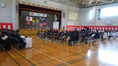 ステージ上に「祝 入学」と書かれた横断幕、国旗と校旗の2つの旗、桜の花びらや子どものイラストが飾られ、壁に紅白幕が貼られた体育館の左側に来賓者、右側に先生方が座り、中央に新入生と保護者が並んで座り、檀上で挨拶を行っている市長の話を参加者が聞いている千徳小学校の入学式の様子の写真