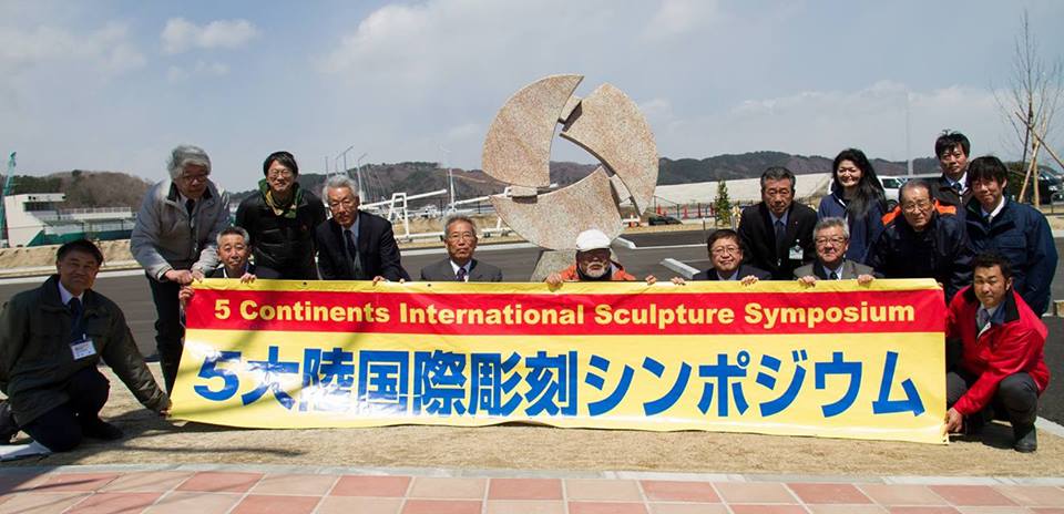 宮古運動公園に寄贈いただいたキャンディス・ギャレット様作「未来ー友情・希望・誠実」と題した、3つの牙の様な形をした物が輪の形を作っている復興支援彫刻が設置された前方に「5大陸国際彫刻シンポジウム」と書かれた横断幕を持って座った市長や14名の関係者の方々が並んでいる記念写真