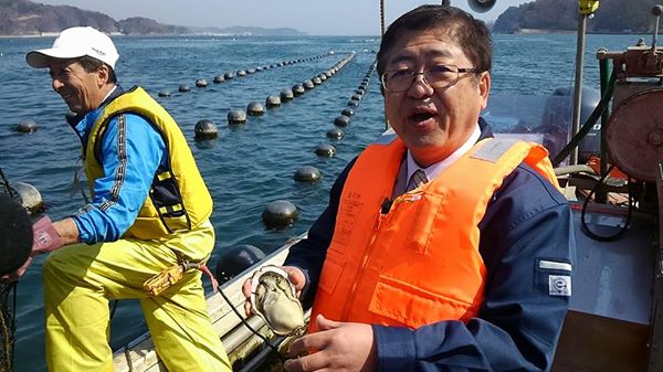 船の上で黄色のライフジャケットを着た男性スタッフの隣で、オレンジ色のライフジャケットを着用した市長が殻を割った「花見かき」を持っている写真