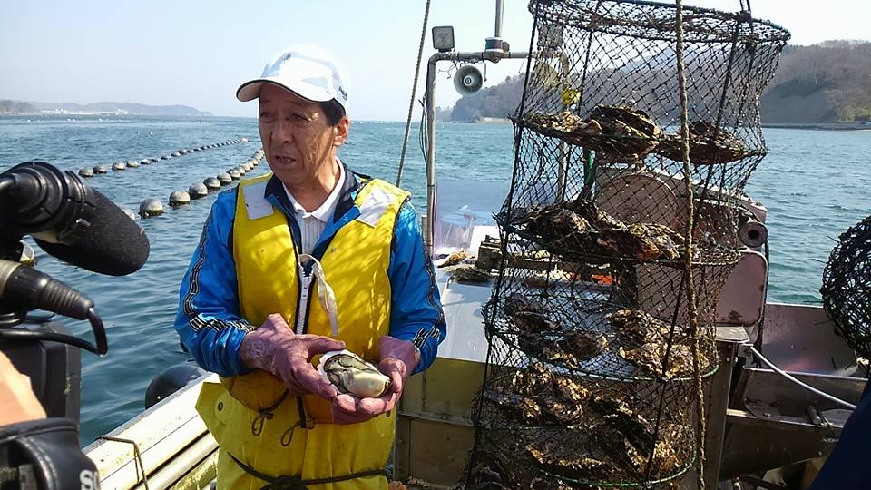 干し網に入った水揚げした大きな「花見かき」の隣で取材を受けている男性スタッフの写真