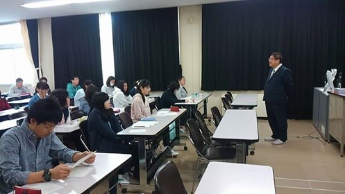 地域総合講座の会場の教室の席に学生が座り、向かい合って立っている市長が話をしている様子の写真