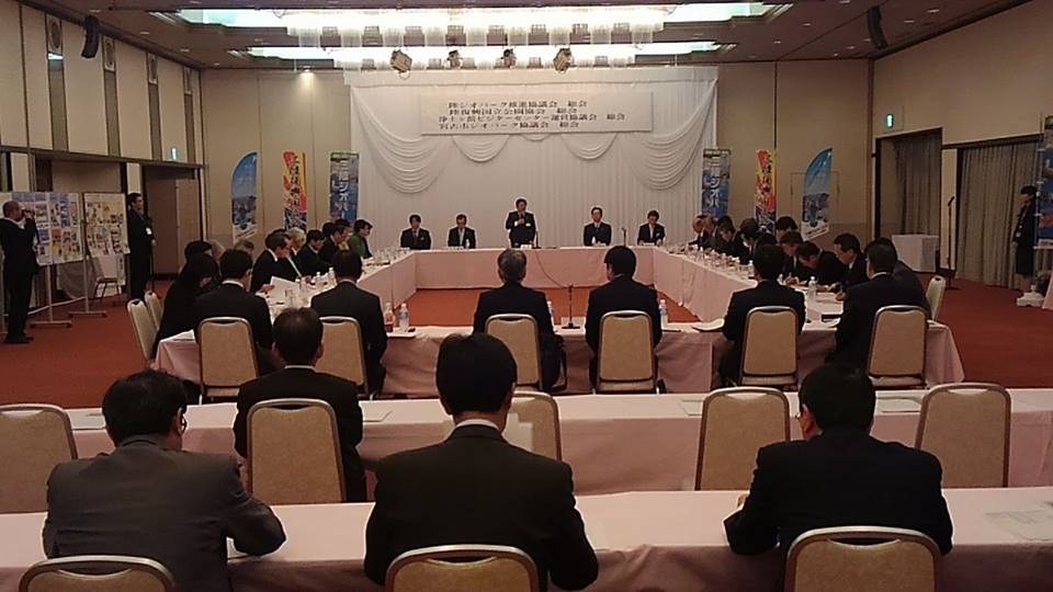 前方に横断幕が掛けられ左右に3つずつ幟旗が設置された会場に白いテーブルクロスが掛けられたロの字型に設置された机と、後方に2列に設置された机があり、前方中央の席から立ち上がった市長が挨拶を行っている写真