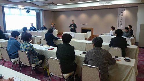 前方左右の関係者と参加者が向かい合って座った会場で、前方中央の席から立ち上がった市長が挨拶を行っている写真