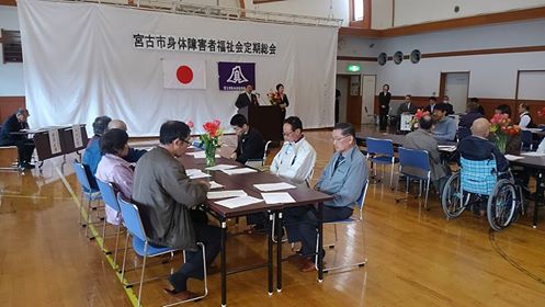 前方に設置された大きな白い幕に「宮古市身体障害者福祉会定期総会」と書かれた横幕と2つの旗が貼られ、グループごとに着席した参加者が、檀上に立つ市長の話を聞いている写真