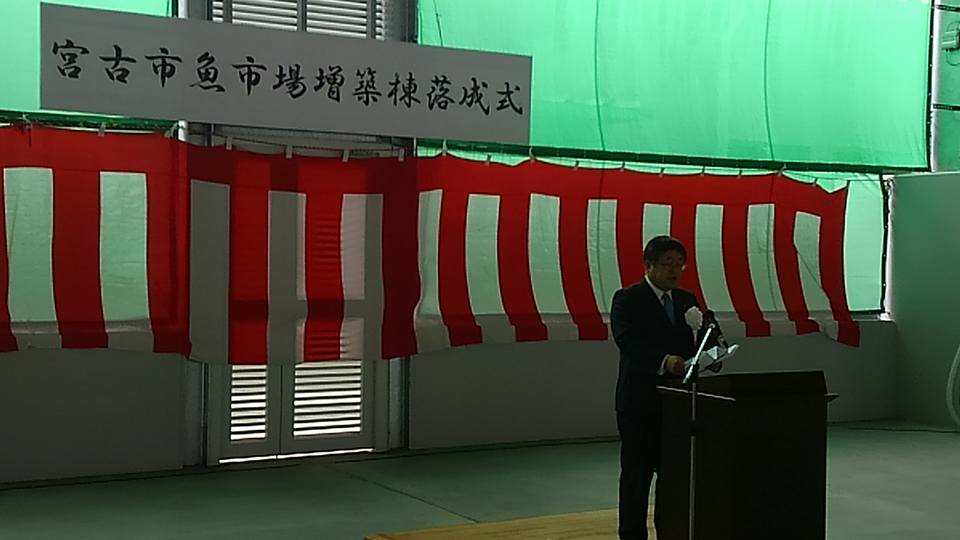 「宮古市魚市場増築棟落成式」と書かれた横断幕と紅白幕が貼られた屋内で、檀上の前に立ち挨拶を行っている市長の写真