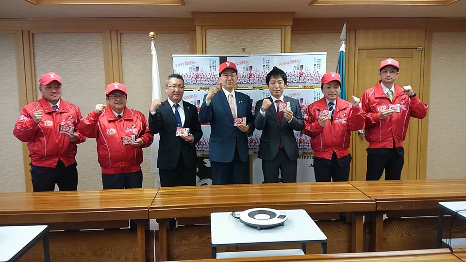 中央に赤色のキャップを被った達増知事とスーツ姿の2名の関係者が立ち、左右にお揃いの赤色のキャップにジャンパーを着用した市長を含めた4名の関係者がガッツポーズをして岩泉ヨーグルト味のキットカットを持って並んでいる写真