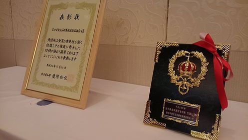 白幕が掛けられた机の上に、額に入った賞状と盾が並んで置かれている写真