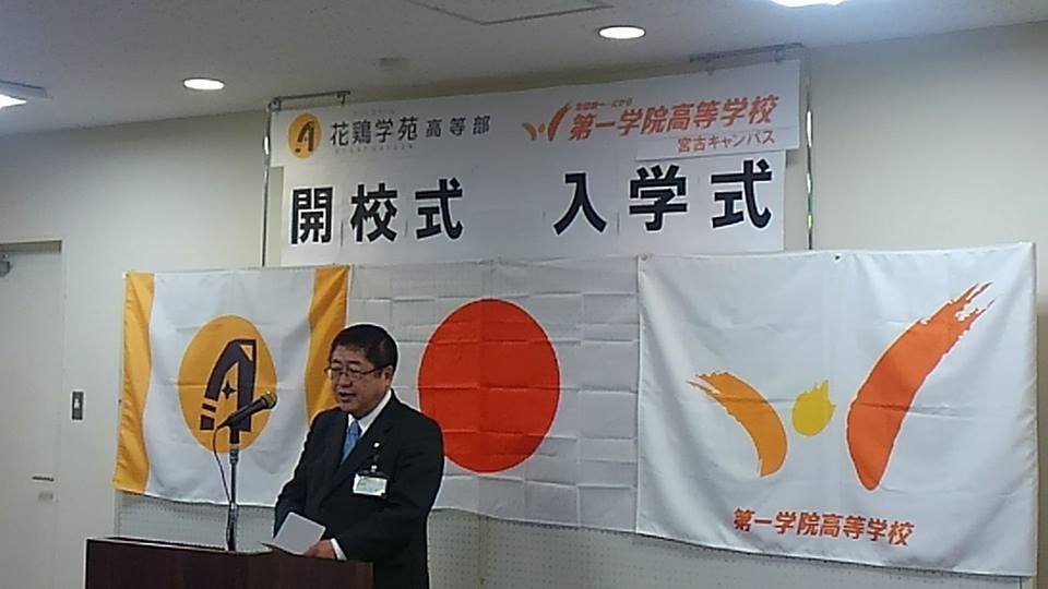 「花鶏学苑高等部開校式 第一学院高等学校宮古キャンパス入学式」と書かれた横幕と3つの旗が壁に貼られた会場の檀上で、市長が挨拶を行っている写真