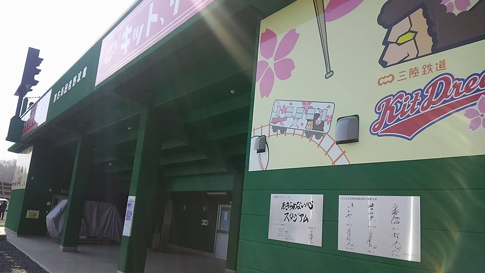 緑色の壁に野球ボールやバット、桜の花びらや鉄道が描かれたイラストと、白地に黒文字で「あきらめない心スタジアム」とサインが書かれている田老野球場の写真