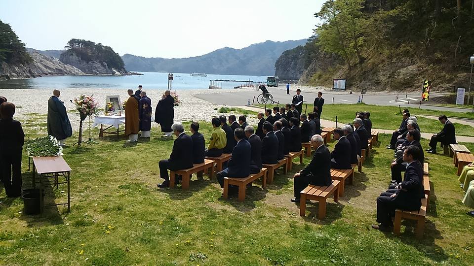 屋外に設置された椅子に礼服を着た大勢の参加者が座り、小さな祭壇の周りに4名のお坊さんがお経を唱えている霊鏡祭の様子の写真