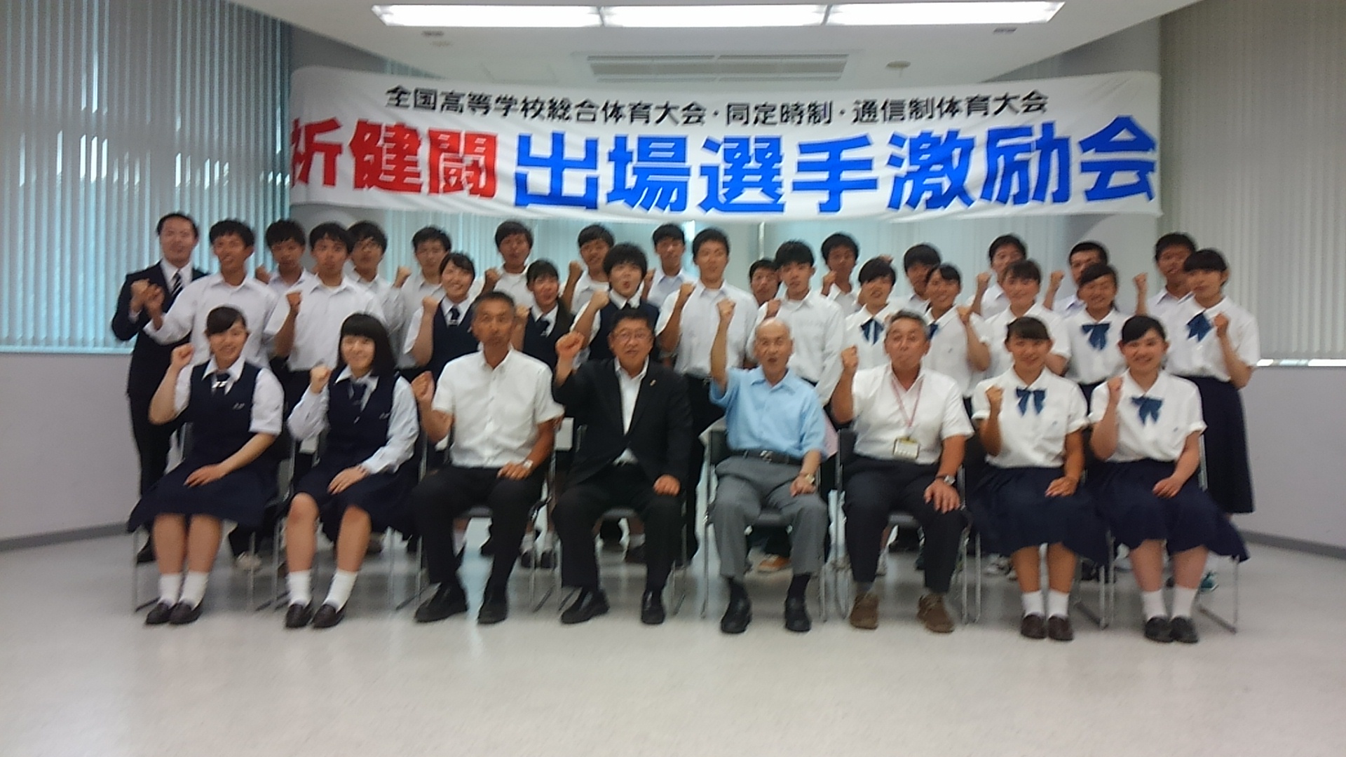 「祈健闘 出場選手激励会」と書かれた横断幕が掛けられた会場で、市長と関係者の男性3名、制服を着た女子高生4名が1列目で椅子に座り、2列目、3列目に立つ高校生たちが並び握った拳を顔の横に掲げてポーズをとっている記念写真