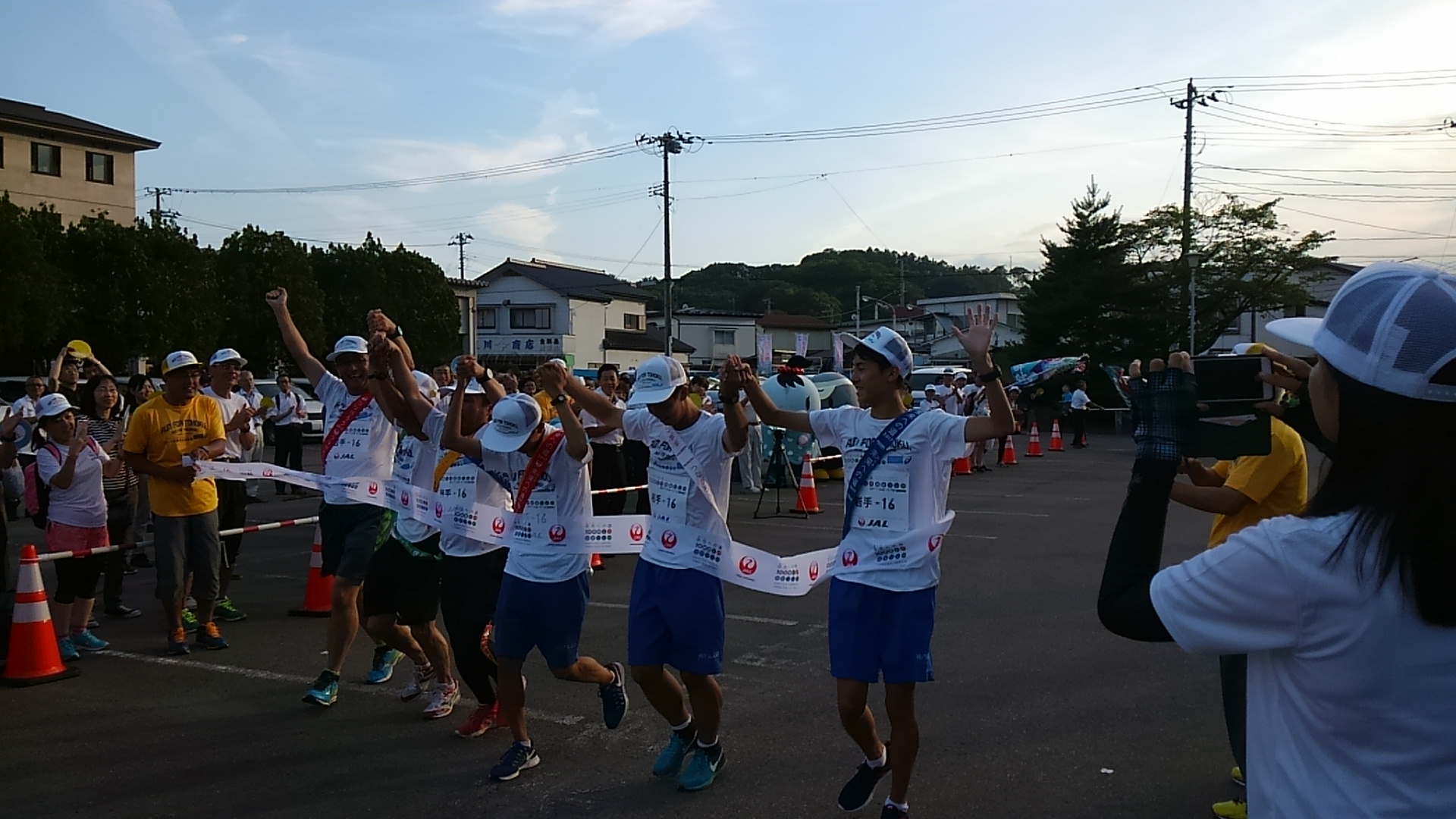 つないだ手をあげながら横一列に並んでゴールテープを切るそれぞれ襷をかけた6名の参加者の男性たちの写真