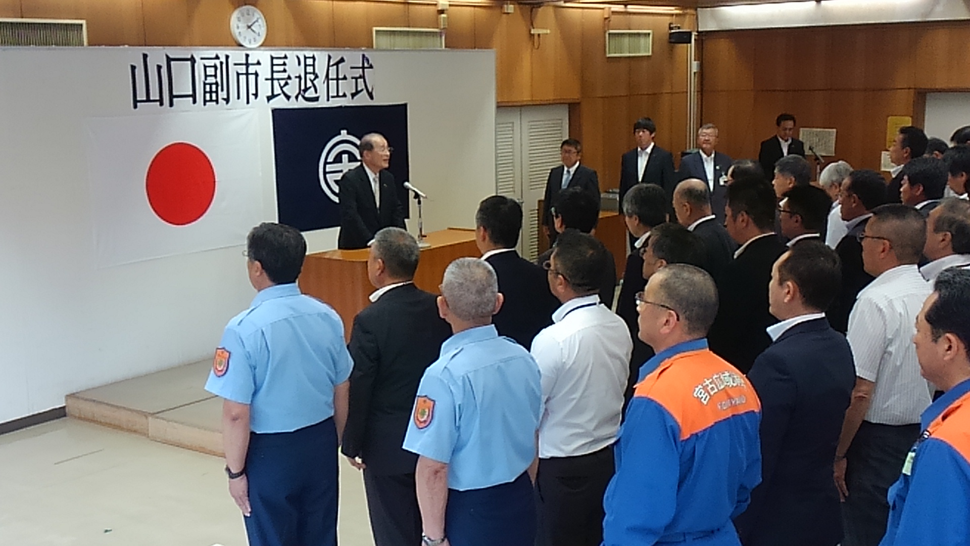 整列する職員の方々の前で、「山口副市長退任式」と書かれた紙と日本国旗、宮古市の市町村旗が貼られたパネルを背に立って話をする山口公正副市長の写真
