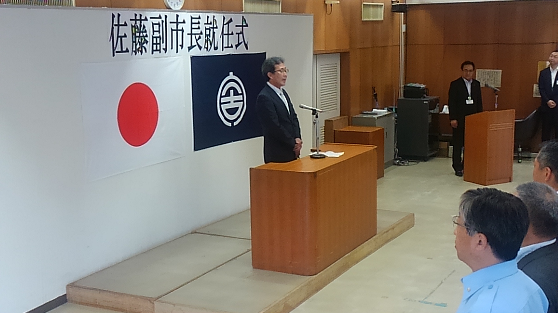 「佐藤副市長就任式」と書かれた紙と日本国旗、宮古市の市町村旗が貼られたパネルを背に、演台に置かれたマイクに向かい話をする様子の佐藤廣昭副市長の写真