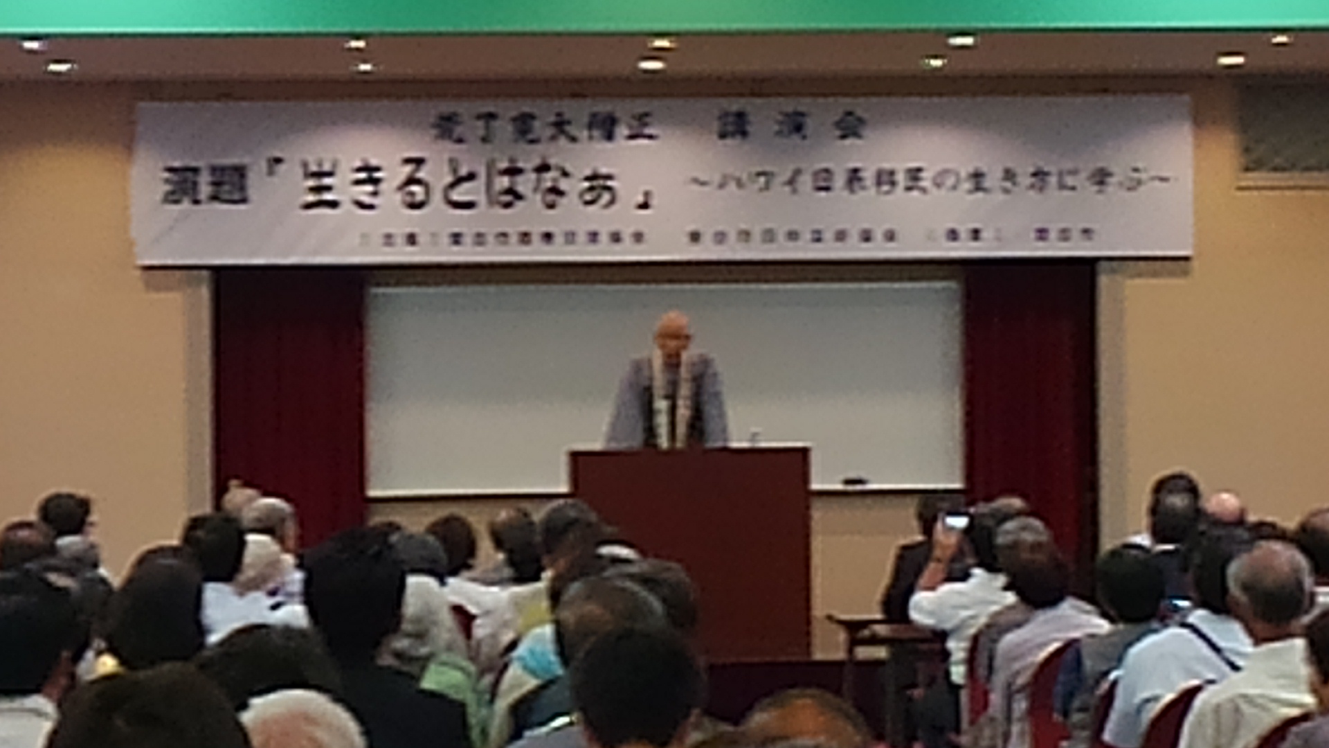 講演会の題名が書かれた横断幕の下に立つ天台宗ハワイ開教総長 荒了寛大僧正と椅子に座り話を聞く様子の参加者たちの写真