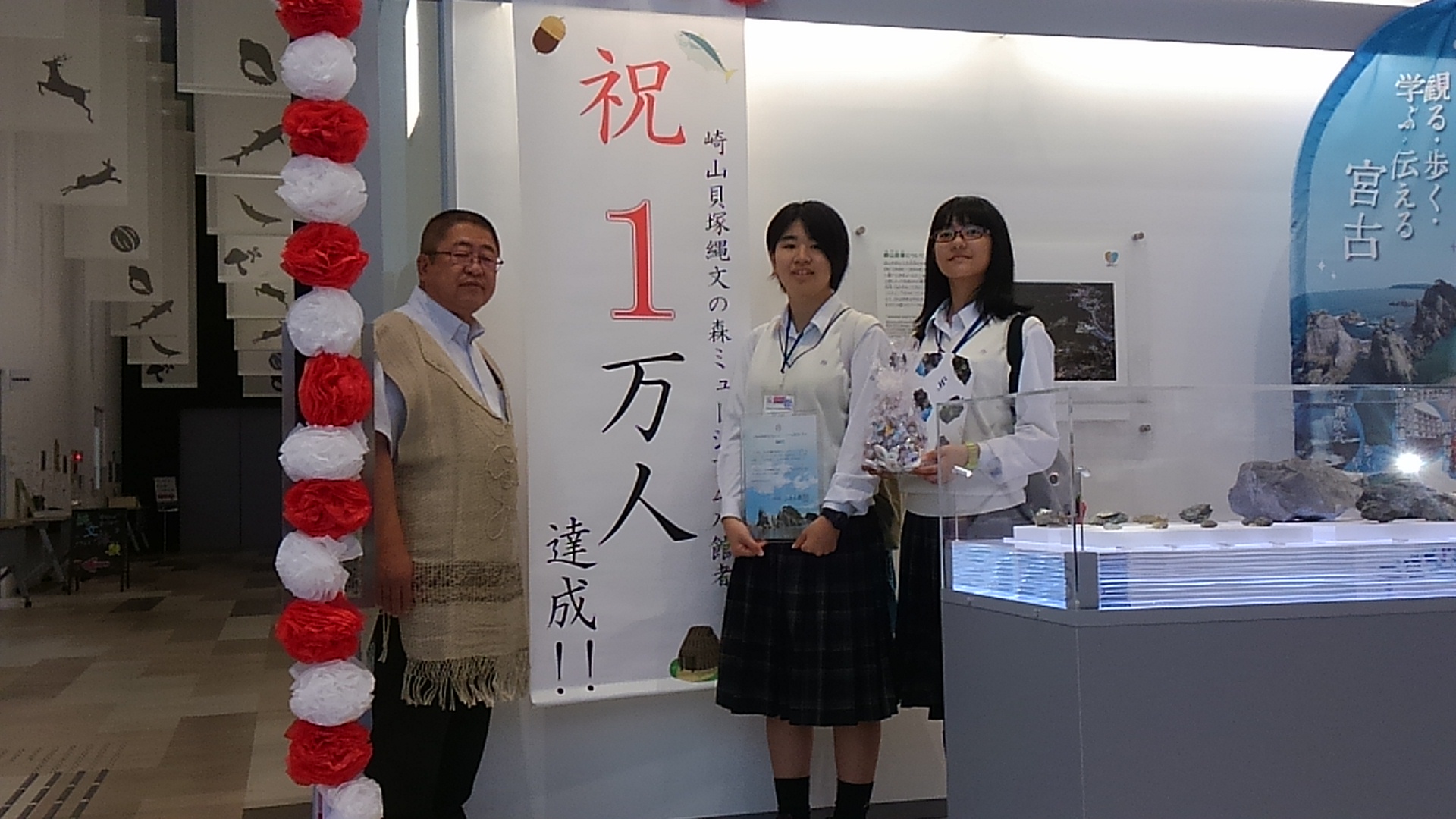 「祝 1万人達成！！」と書かれた垂れ幕の左側に立つ市長と、右側に立つ2名の制服を着た女子中学生の記念写真