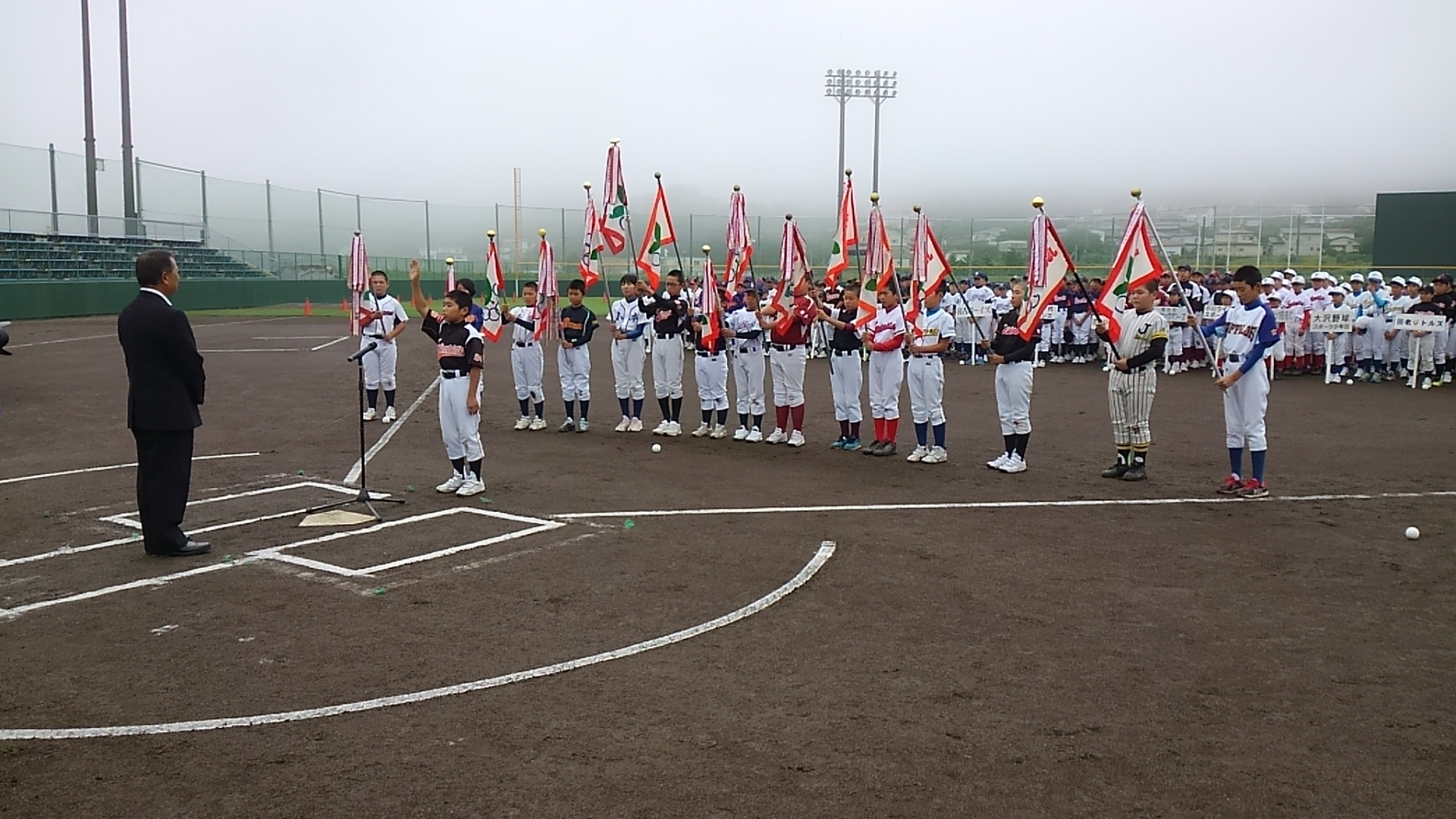 野球場のホームベースに置かれたスタンドマイクに向かい、右手を上にあげて宣誓する男の子と、その後方で旗を掲げる15名の選手たちの写真