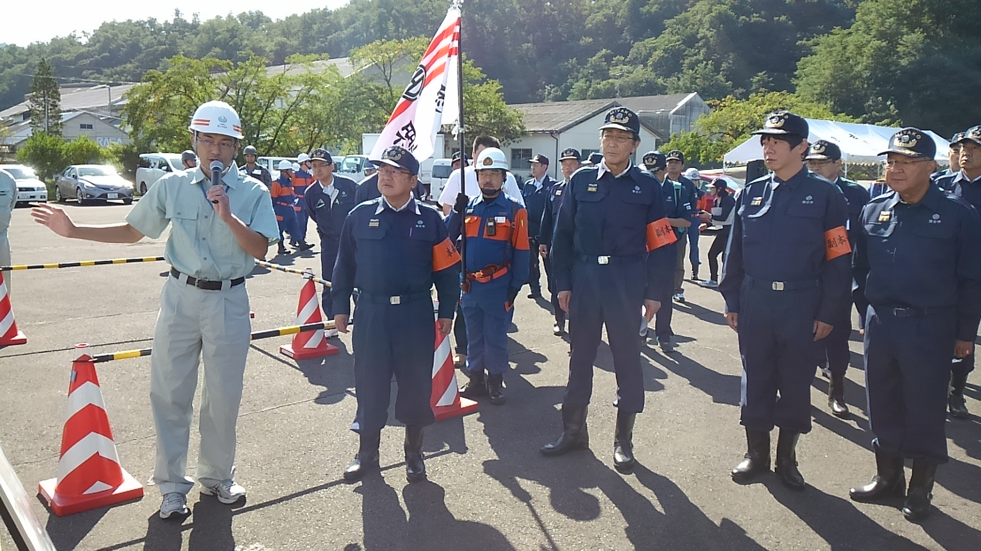 紺色の作業服を着てオレンジ色の腕章を着けた市長と関係者の男性たちが、マイクを持つ男性の話を聞いている様子の写真