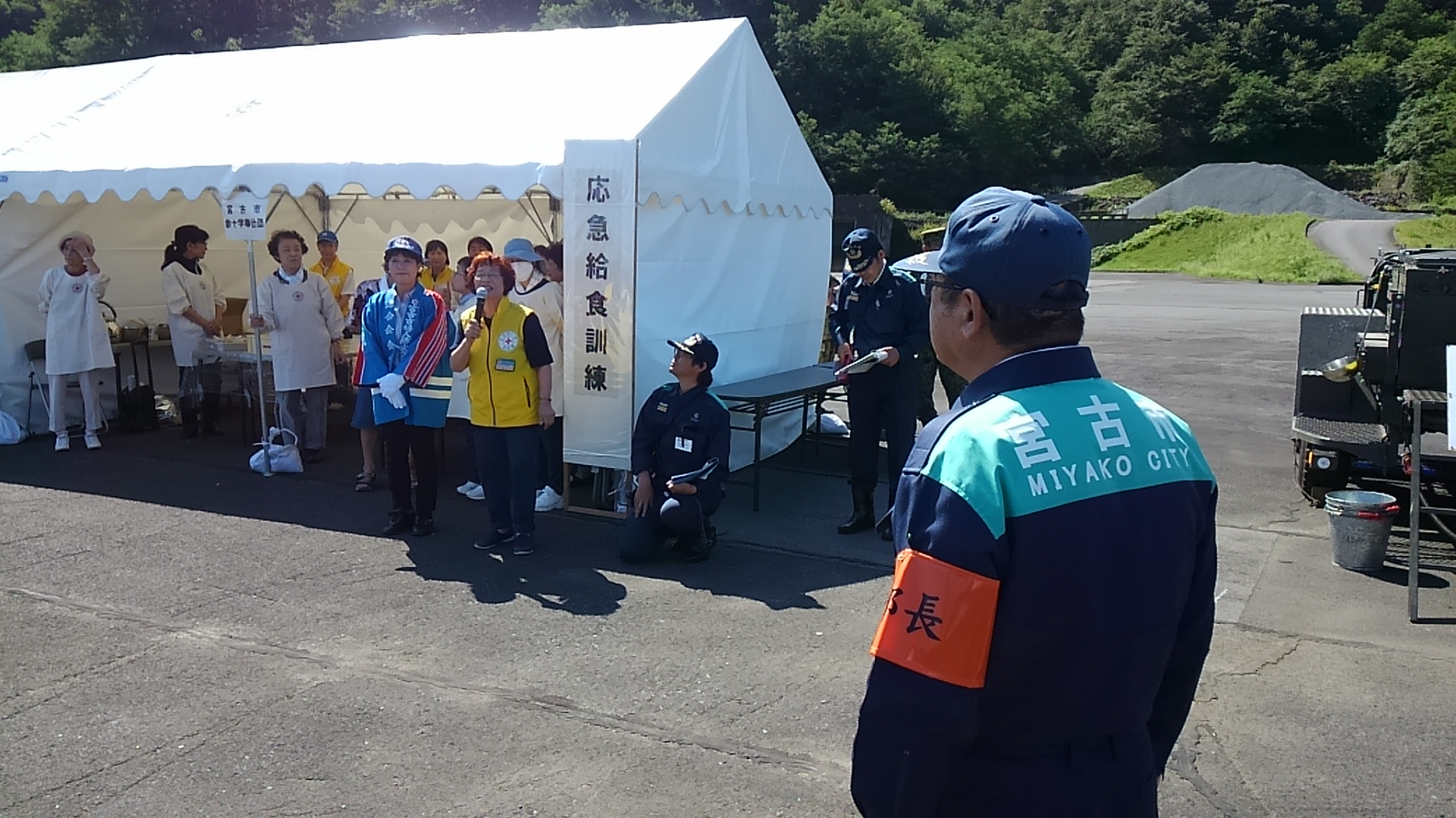紺色の作業服を着てオレンジ色の腕章をつけた市長が、白いテントの前で話をする黄色いベストを着た女性の方を見ている写真