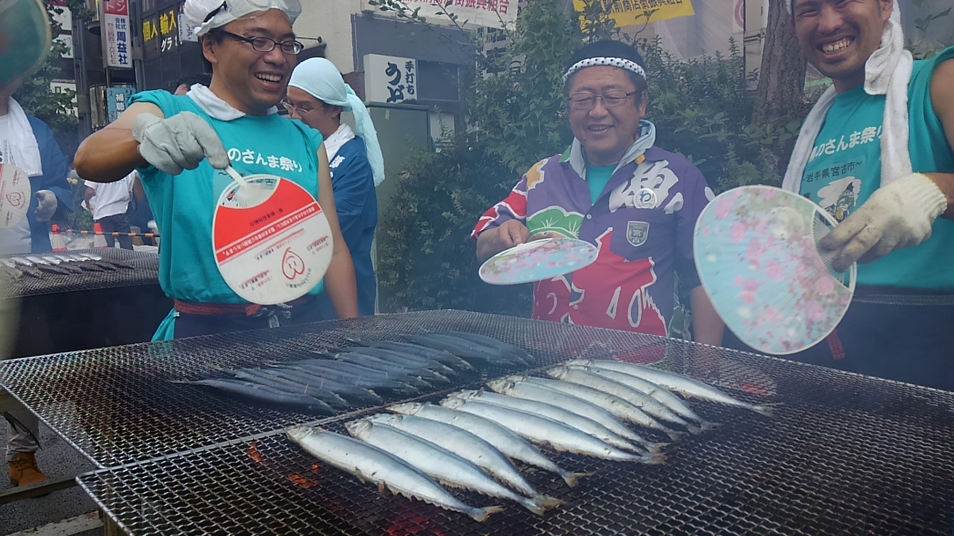 大漁旗で作られたシャツを着た市長と水色のシャツを着た2名の男性スタッフが、網の上に並べたサンマをうちわで扇いでいる目黒のさんま祭りの様子の写真