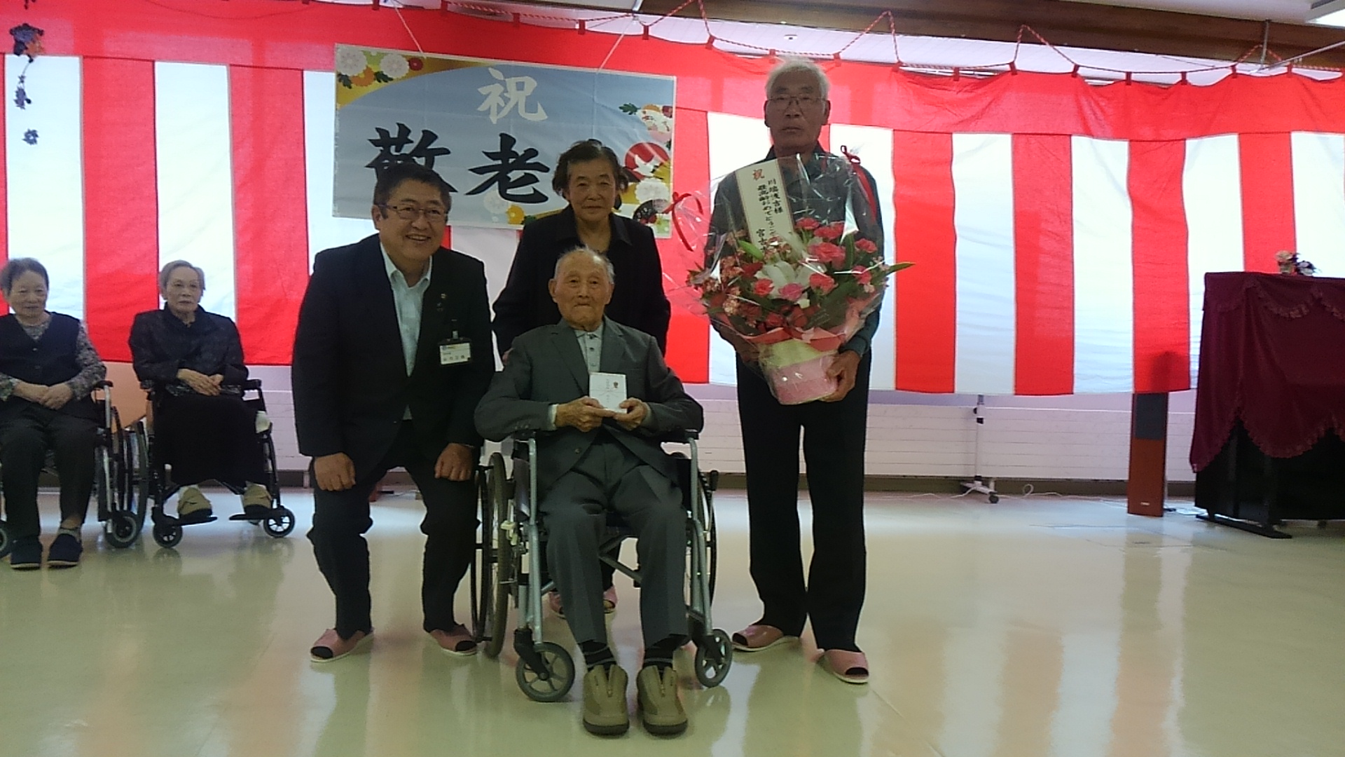 後方に紅白幕が飾られた介護老人保健施設「桜ヶ丘」で、車いすに座った市内最高齢の川端浅吉様の後ろにお祝いの花を持ったご家族と市長が並んでいる記念写真