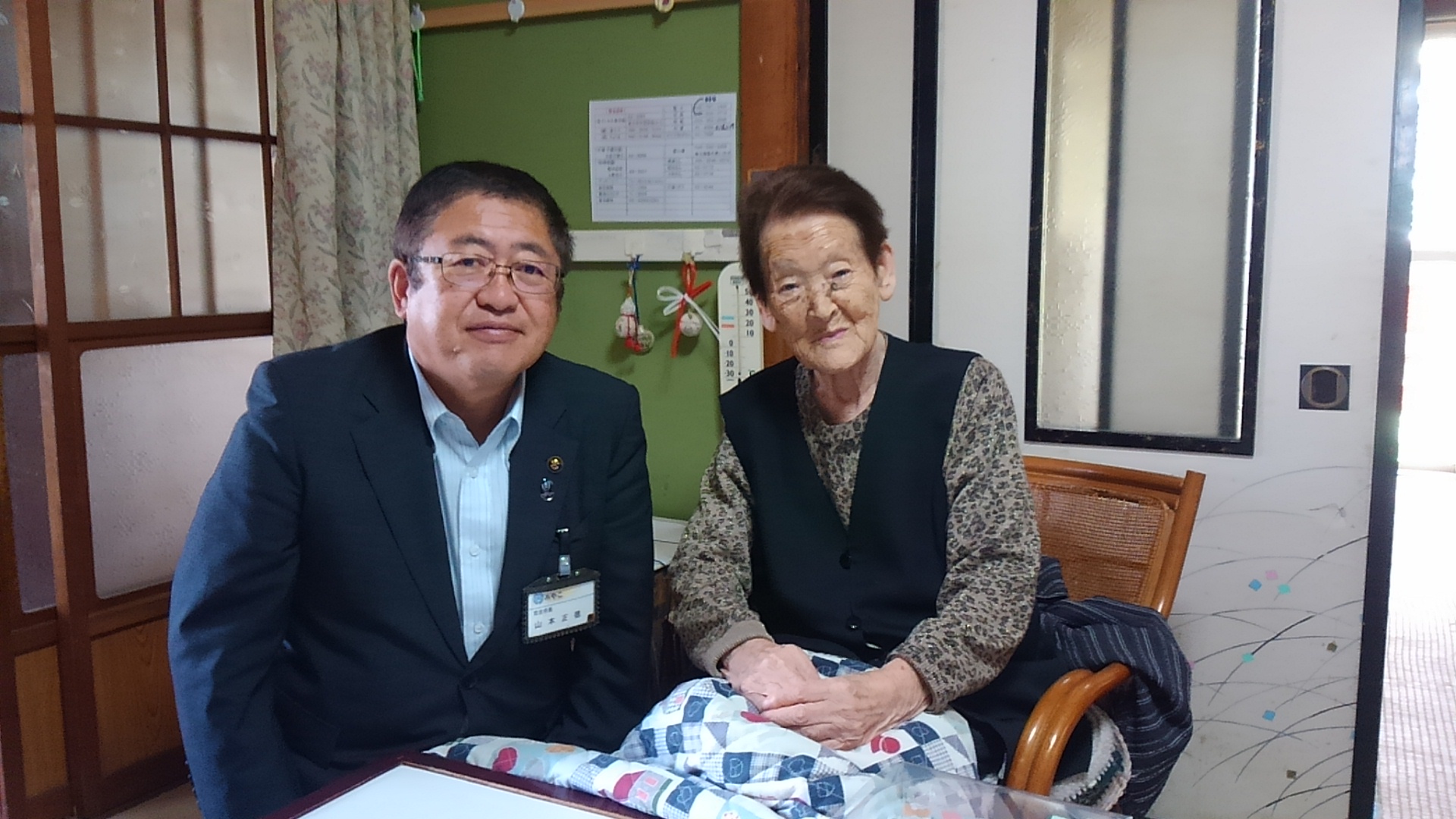 コタツが置かれたリビングで、藤の椅子に座った大久保貞子様と市長が並んで座っている百歳祝いの記念写真