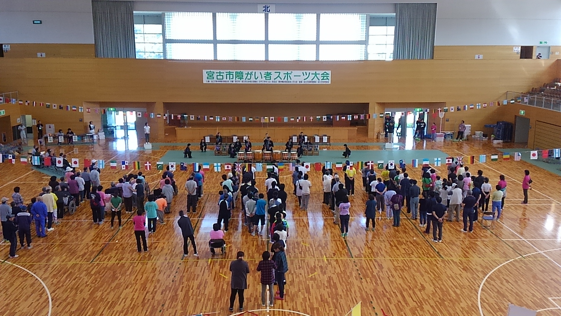 壁に「宮古市障がい者スポーツ大会」と書かれた横幕が貼られ、様々な国の国旗が飾られた体育館に参加者が整列し、前方で宮古商業高校の皆さんによる海鳴太鼓が披露されている様子の写真