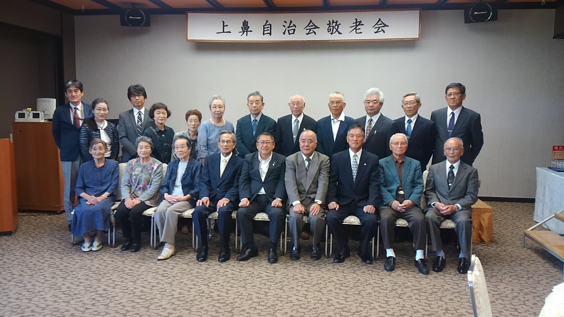 「上鼻自治会敬老会」と書かれた横断幕が掲げられた会場で、前列の椅子に市長と8名、後列に10名の参加者と2名の関係者が並んでいる集合写真