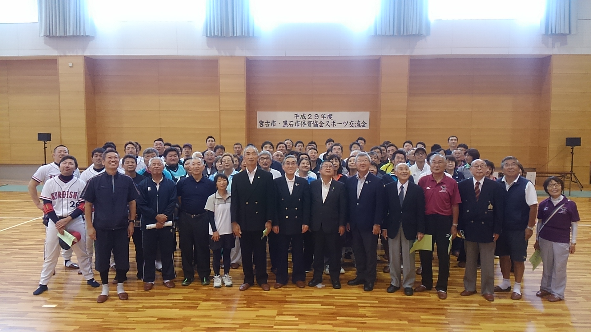 「平成29年度 宮古市・黒石市体育協会スポーツ交流会」と書かれた横幕が体育館の壁に貼られ、市長を中央に関係者や参加者が並んで上を見ている集合写真