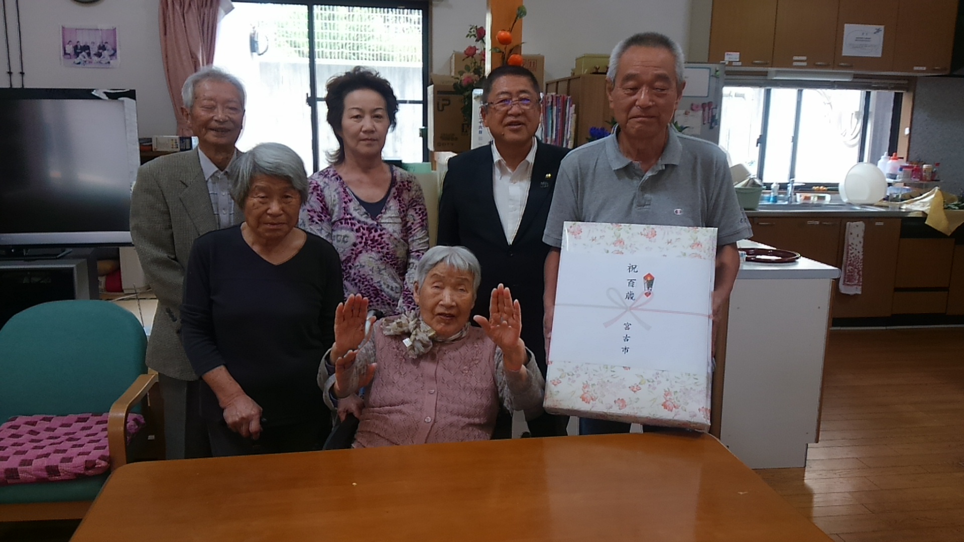 「祝 百歳 宮古市」と書かれた熨斗紙がかけられたお祝いの品を男性が持ち、真ん中の椅子に座り両手を挙げた藤田チエ子様と4名のご家族と市長が並んでいる百歳祝いの記念写真
