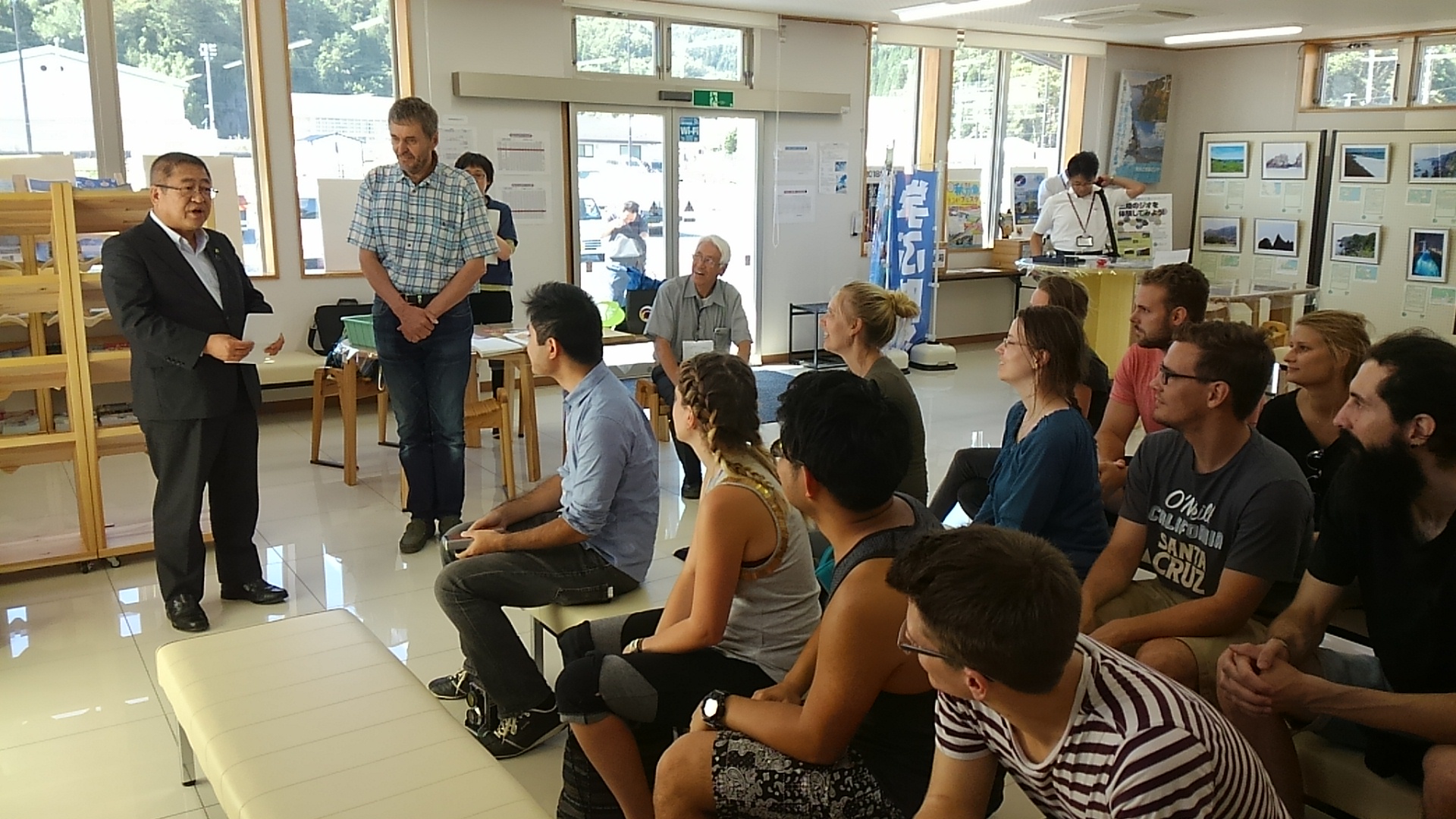 3列に並んだ長椅子にサマースクールに訪れた15名のブラウンシュヴァイク工科大学生やが座り、右側に立つ市長の話を熱心に聞いている写真
