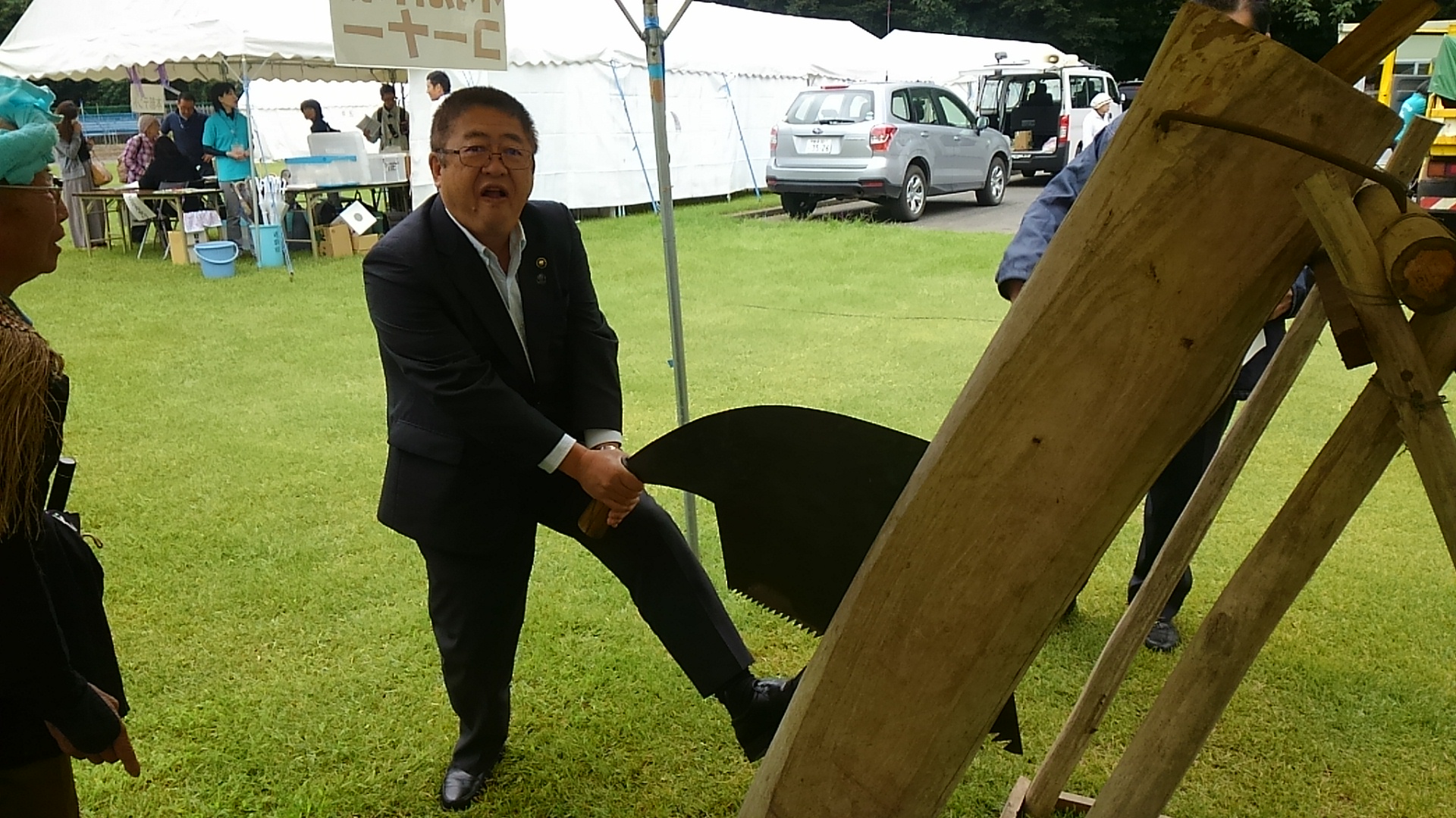 やまびこフェスタ会場のブースで、大きなのこぎりを両手で持ち大変そうに木材を切っている市長の写真